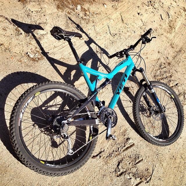 Yeti 575: A turquoise mountain bike placed on sandy terrain, showcasing its sleek frame and sturdy wheels, with the brand name "Yeti" visible on the frame. The bike is positioned to highlight its various components, including the handlebars and drivetrain.