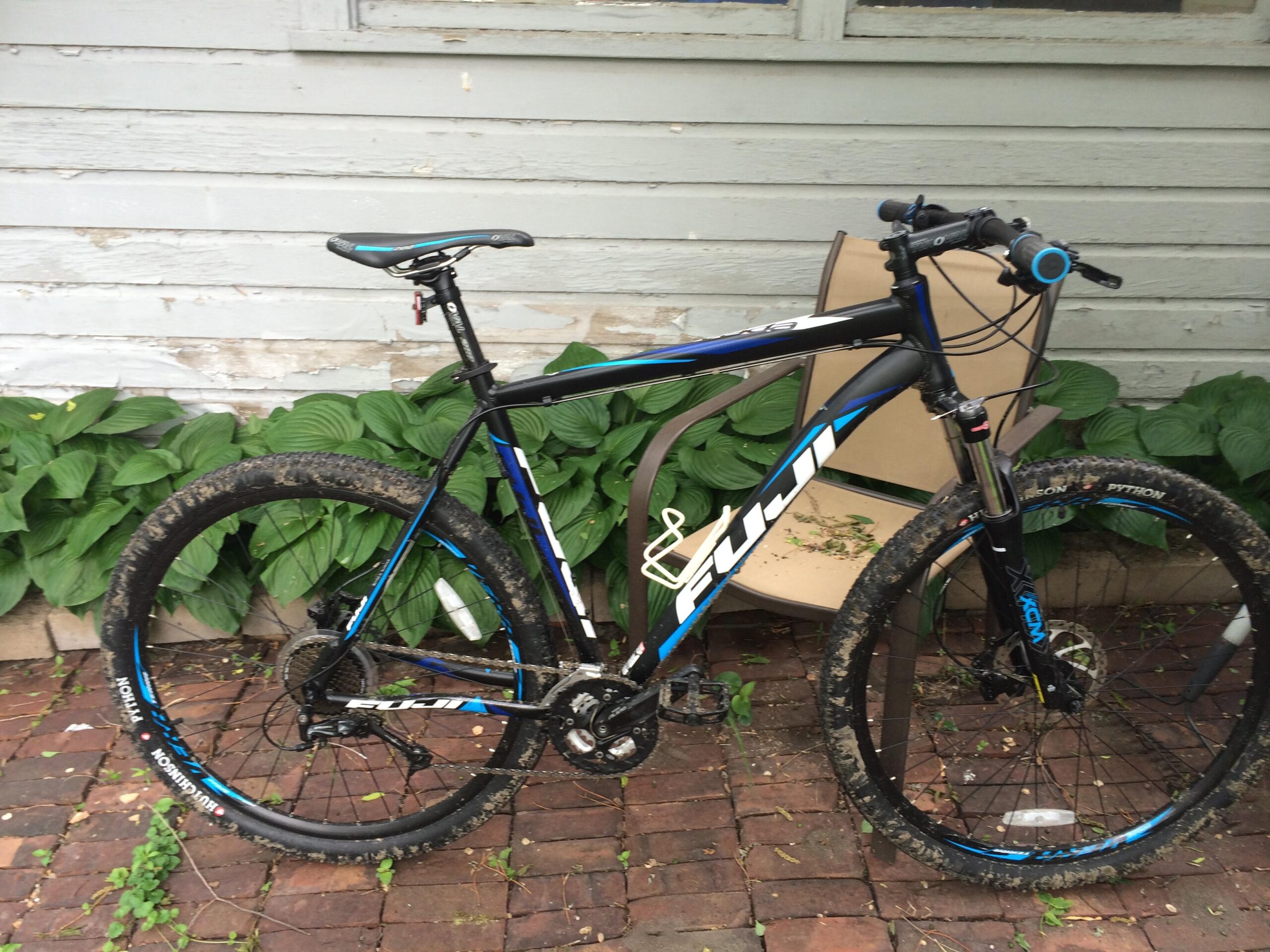 Fuji nevada 1.5 29er: A black and blue mountain bike with thick, muddy tires is parked next to a beige chair in front of a gray wooden wall. Lush green hostas are visible in the background, and the bike shows signs of use, particularly on the tires.