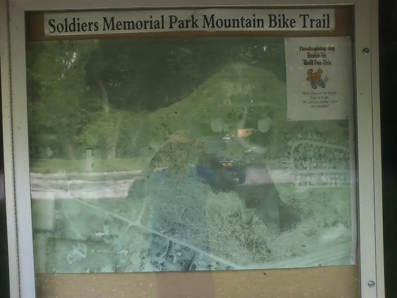 A weathered informational sign for Soldiers Memorial Park Mountain Bike Trail, displaying a faded map of the trail and park layout. The sign includes a notice for a Thanksgiving Day event with details about meeting at a kiosk. The background features greenery and a glimpse of nearby paths. Soldiers Memorial Park Bike Trail mountain bike trail.