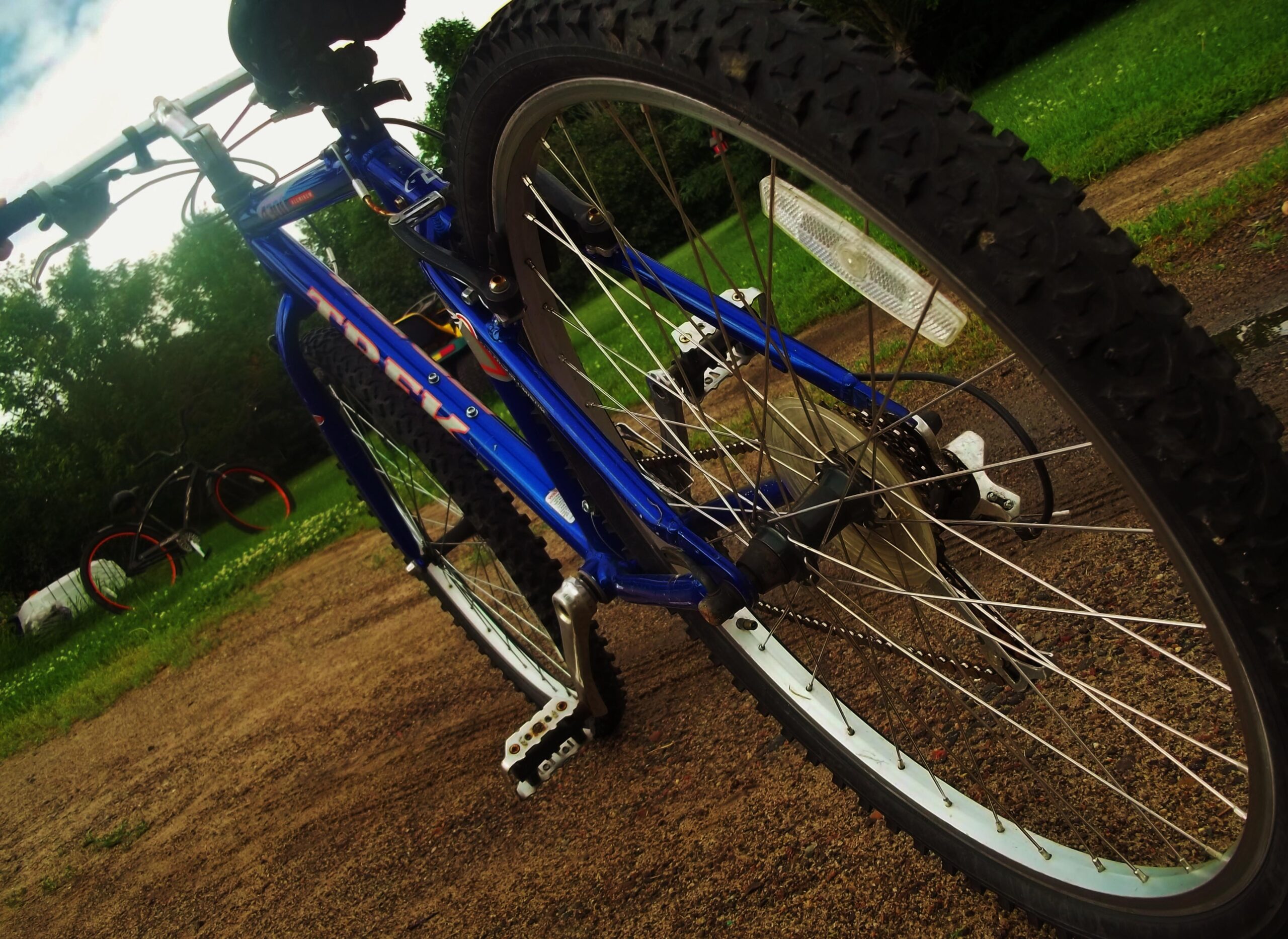 Trek 4300: A close-up view of a blue mountain bike positioned at an angle, showing its rear wheel, tire, and chain, with greenery and another bike in the background on a gravel path.