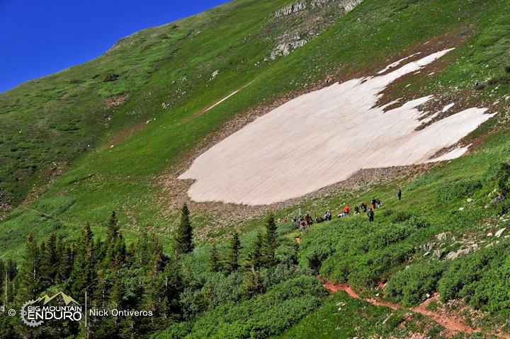 A landscape photograph showing a vibrant green mountainside with a prominent patch of remaining snow. In the foreground, a group of people is gathered, likely enjoying outdoor activities. The scene captures the beauty of nature, highlighting both the lush vegetation and the snow-covered area, set against a clear blue sky. Colorado Trail: Kennebec Pass To Junction Creek mountain bike trail.