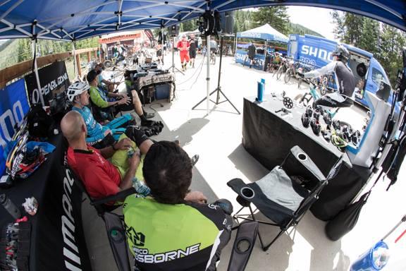 A group of cyclists and support staff seated under a blue tent at a biking event, with a view of the event area showcasing bicycles, equipment, and vendors. The scene captures the camaraderie and preparation atmosphere before a race. Keystone Resort Bike Park mountain bike trail.