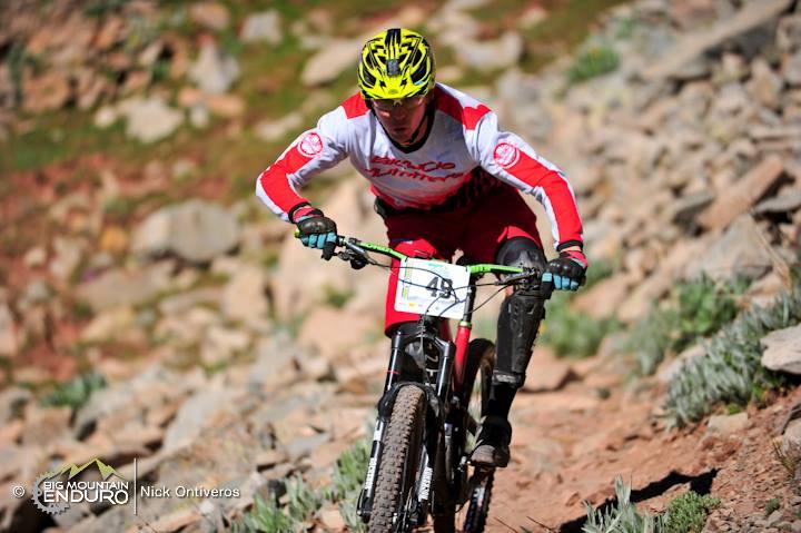 A mountain biker in a red and white jersey navigates a rocky trail, wearing a bright yellow helmet. The rider appears focused and determined, showcasing a dynamic pose as they maneuver down the slope. Green vegetation and rugged terrain surround the scene, highlighting the outdoor setting. Colorado Trail: Kennebec Pass To Junction Creek mountain bike trail.