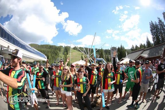A group of enthusiastic participants wearing colorful jerseys in red, green, and yellow gather outdoors, smiling and posing for the camera. The background features a ski lift and green hills under a bright blue sky with fluffy white clouds. Tents and equipment are set up nearby, indicating a festive atmosphere. Keystone Resort Bike Park mountain bike trail.