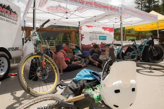 A group of four individuals resting on the ground near a bike recovery station, surrounded by various bicycles and gear. A mountain bike with a yellow wheel is in the foreground, and a tent overhead provides shade. The setting suggests an outdoor cycling event, with equipment and a partially visible banner in the background. Keystone Resort Bike Park mountain bike trail.