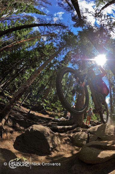 A mountain biker in mid-air jumps over a rocky obstacle in a forested area, with sunlight filtering through the trees above. The scene captures the thrill of mountain biking, surrounded by tall trees and a clear blue sky. Keystone Resort Bike Park mountain bike trail.