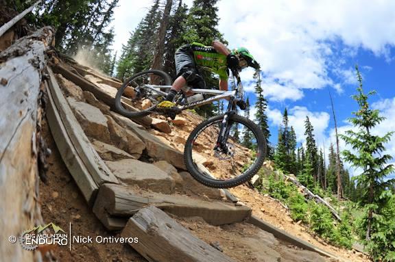 A mountain biker navigating a rocky downhill trail in a forested area, with sunlight filtering through the trees and a blue sky dotted with clouds. The rider is positioned mid-action, leaning forward on a mountain bike, while large boulders and wooden planks are visible along the path. Keystone Resort Bike Park mountain bike trail.