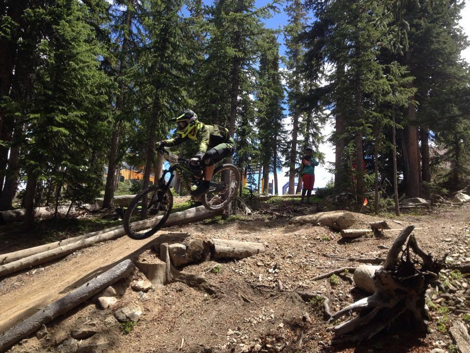 A mountain biker in a green and black outfit jumps over a small log on a dirt trail surrounded by tall pine trees. In the background, another person watches and captures the moment with a camera. The sky is bright blue with a few clouds. Keystone Resort Bike Park mountain bike trail.