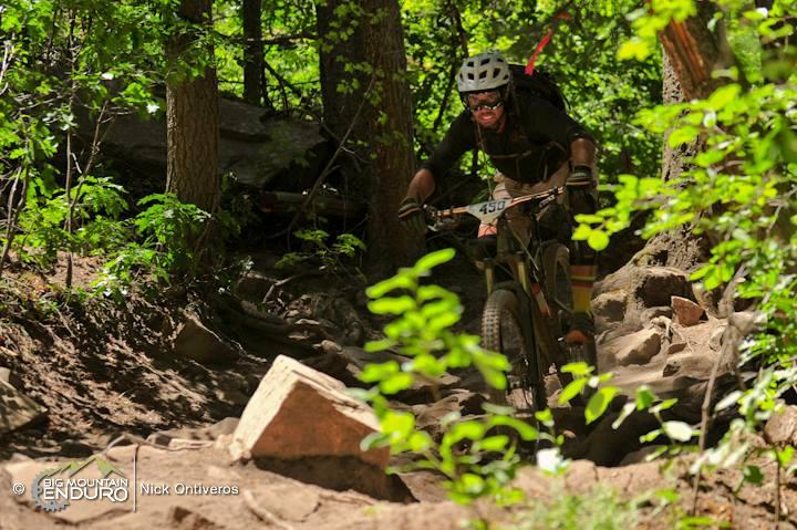 A mountain biker navigating a rocky trail through a lush green forest, focused and determined, with sun filtering through the trees. The cyclist is wearing a helmet and biking gear, with the number 450 visible on their bike. Colorado Trail: Kennebec Pass To Junction Creek mountain bike trail.