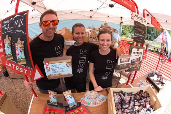 Three enthusiastic individuals in black t-shirts are standing behind a table at an outdoor event. The man on the left, wearing sunglasses, holds a sign that reads "Latest Trail Mix." The two women beside him smile and display signs for "CLIF Bar" and "Energize." A variety of energy bars and snacks are arranged on the table, along with promotional materials in a festive, tented setting. Keystone Resort Bike Park mountain bike trail.