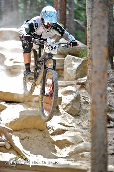 A mountain biker in action, leaping off a rocky incline amidst a forested background. The rider is wearing protective gear, including a helmet and gloves, and has a race number displayed on their bike. Dust is kicked up around them as they navigate the rugged terrain. Keystone Resort Bike Park mountain bike trail.