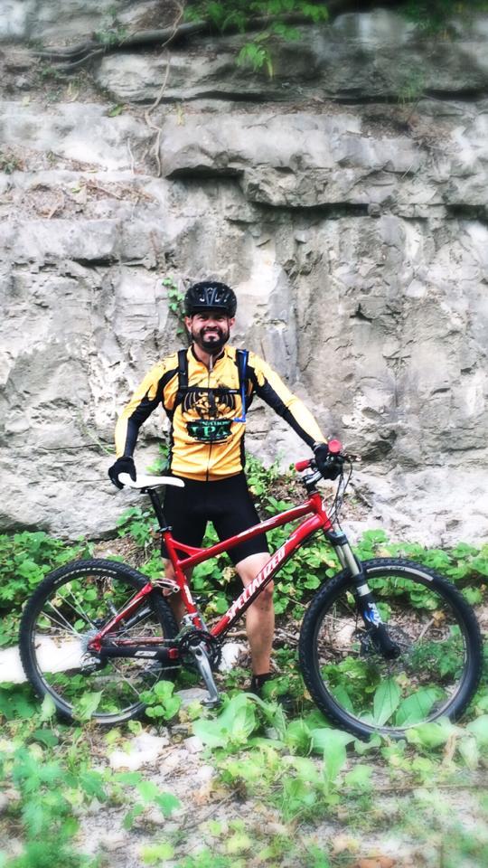 Specialized Epic Expert: A person in cycling gear stands next to a red mountain bike, with a rocky wall in the background. The cyclist wears a yellow and black jersey and a helmet, surrounded by greenery and vegetation.