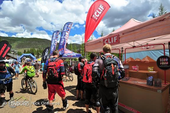 A group of mountain bikers in casual cycling attire stand in line at a vendor booth during an outdoor event. Colorful flags and banners from various sponsors surround the area, while tents are set up in the background against a backdrop of green trees and a partly cloudy sky. The scene captures a lively atmosphere typical of biking festivals. Keystone Resort Bike Park mountain bike trail.