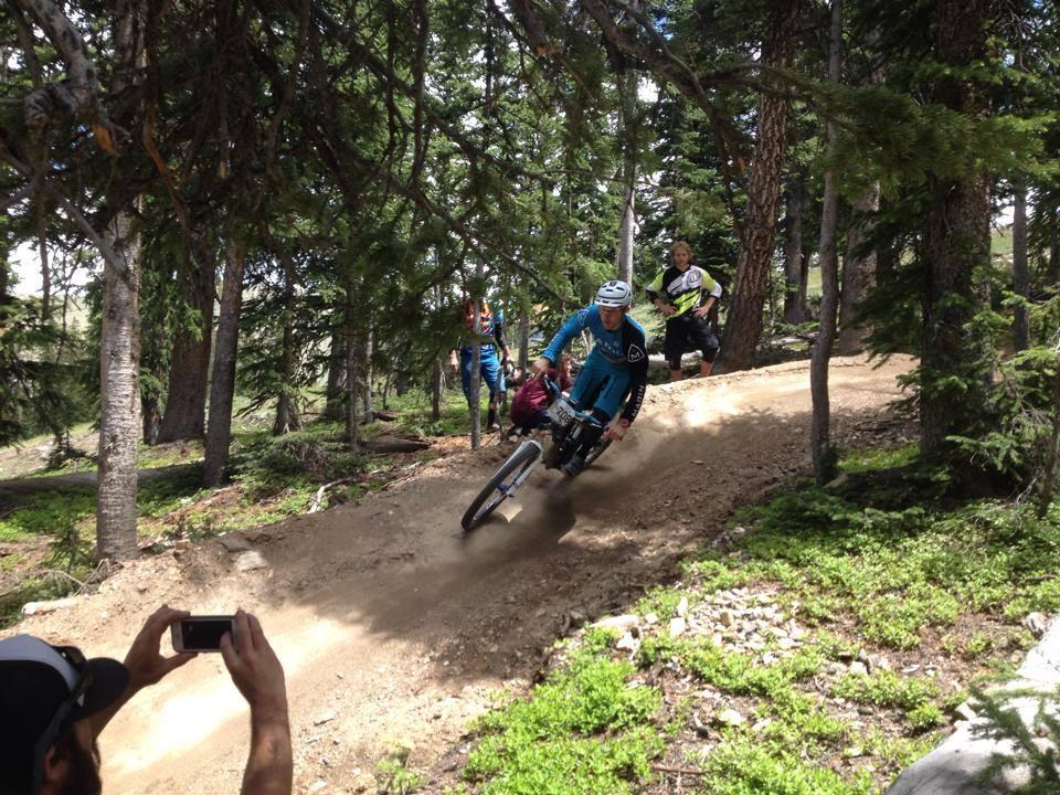 A mountain biker in a blue jersey flies around a curve on a dirt trail surrounded by trees, kicking up dust behind them. In the background, two other riders watch and a person captures the moment with a smartphone. The scene depicts an exciting outdoor biking adventure in a forested area. Keystone Resort Bike Park mountain bike trail.