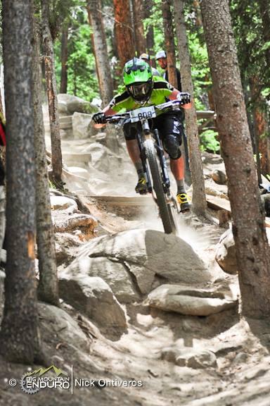A mountain biker navigating a rocky terrain surrounded by tall trees, with dust kicked up from the trail. The rider is wearing a green helmet and jersey, focusing on the path ahead. Another rider can be seen in the background. Keystone Resort Bike Park mountain bike trail.