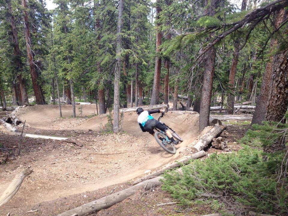 A mountain biker navigating a curving dirt trail through a dense forest, surrounded by tall pine trees and fallen logs. The rider is leaning into the turn, showcasing a dynamic riding position. Keystone Resort Bike Park mountain bike trail.