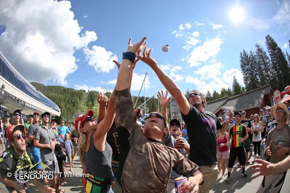 A lively outdoor scene featuring a crowd of people at a festival or event, with some individuals reaching up to grab an object thrown in the air. The background showcases trees and a cloudy blue sky, while attendees are dressed casually and appear to be enjoying themselves, holding drinks and engaging with one another. Keystone Resort Bike Park mountain bike trail.