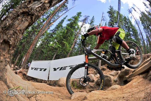 A mountain biker jumps over rocky terrain in a wooded area, wearing a red shirt and lime green shorts. A Yeti brand banner is visible in the background, along with tall trees and a clear blue sky. The image has a fish-eye perspective, emphasizing the action and the environment. Keystone Resort Bike Park mountain bike trail.
