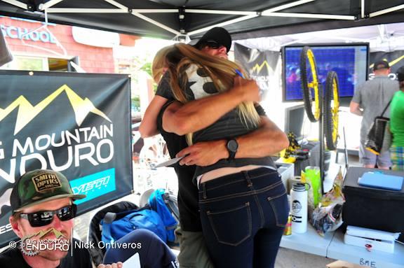 A celebration at a mountain biking event, featuring two people embracing joyfully. One individual is wearing a cap and sunglasses, while the other is in casual attire. The background includes promotional banners for Big Mountain Enduro and various biking equipment, creating a lively atmosphere. Keystone Resort Bike Park mountain bike trail.