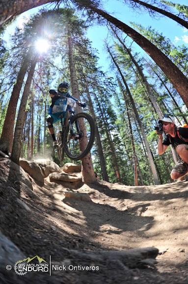 A mountain biker performs a jump over a rocky section of a forest trail, with tall trees in the background. A photographer is capturing the action, positioned nearby. The scene is illuminated by bright sunlight filtering through the trees. Keystone Resort Bike Park mountain bike trail.