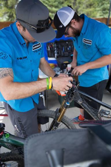 Two mechanics in matching blue shirts work on a mountain bike in a workshop setting. One mechanic is focused on adjusting the bike's suspension components, while the other appears to be assisting or preparing tools in the background. Various bike tools and equipment are visible on a table nearby. The scene captures a busy, collaborative atmosphere typical of bike maintenance. Keystone Resort Bike Park mountain bike trail.