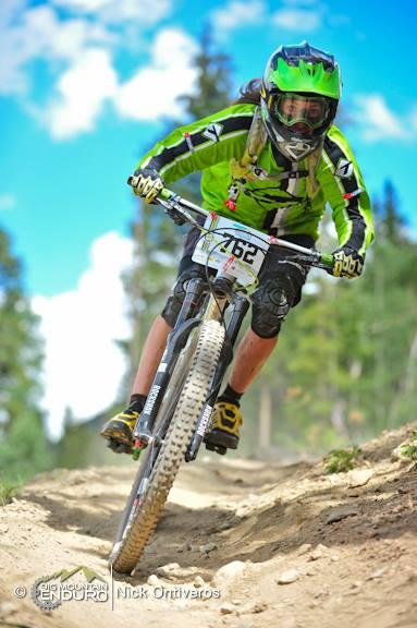 A mountain biker in a bright green outfit navigates a dirt trail, leaning forward as they ride downhill. The rider is wearing a helmet and protective gear, showcasing a focus on safety while racing. The background features trees and a blue sky with fluffy white clouds, indicating an outdoor setting. Keystone Resort Bike Park mountain bike trail.