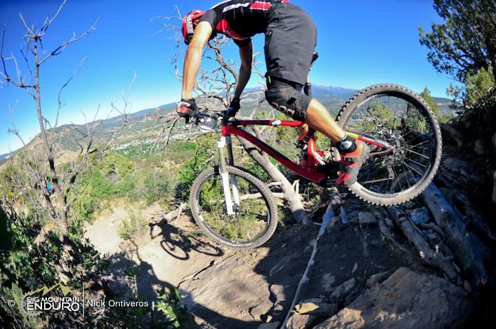 A mountain biker performing a jump on a rocky trail, with a clear blue sky in the background and green hills in the distance. The rider is wearing a helmet, red and black jersey, and shorts, showcasing an action-packed moment in outdoor cycling. Raider Ridge mountain bike trail.