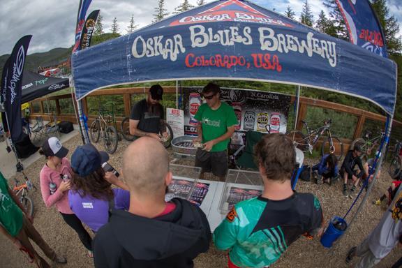 A group of people gathered around a booth with an "Oskar Blues Brewery" banner, engaging in activities related to cycling. Several bicycles are parked nearby, and attendees are interacting with staff at the booth, which features promotional materials. The setting is outdoors, surrounded by trees and natural scenery. Keystone Resort Bike Park mountain bike trail.