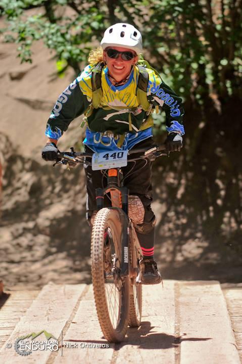 A mountain biker wearing a helmet, sunglasses, and a colorful jersey, riding up a wooden ramp with a smiling expression. The background features greenery, indicating an outdoor setting. The bike has thick tires designed for rough terrain, and the rider's number, 440, is visible on the front. Colorado Trail: Kennebec Pass To Junction Creek mountain bike trail.