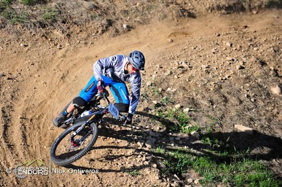 A mountain biker skillfully navigates a dirt trail, leaning into a sharp turn. The rider is wearing a helmet and protective gear, showcasing a dynamic pose while riding a blue mountain bike on a rocky, sunlit terrain. Keystone Resort Bike Park mountain bike trail.