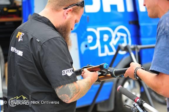 A mechanic with a beard examines a mountain bike fork in a workshop setting, while another person assists by holding the fork. The background features a blue van with the logo "PRO" visible. Keystone Resort Bike Park mountain bike trail.