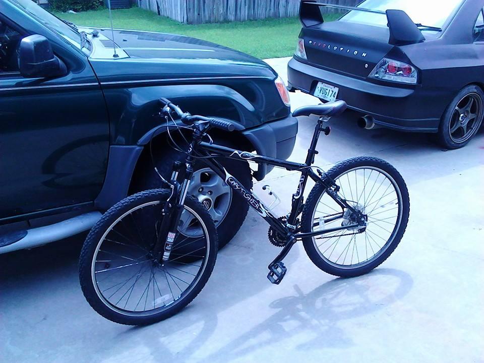 Gary Fisher Wahoo: A black mountain bike parked next to a green SUV in a driveway, with a dark-colored sports car partially visible in the background. The setting features a grassy area and wooden fence.