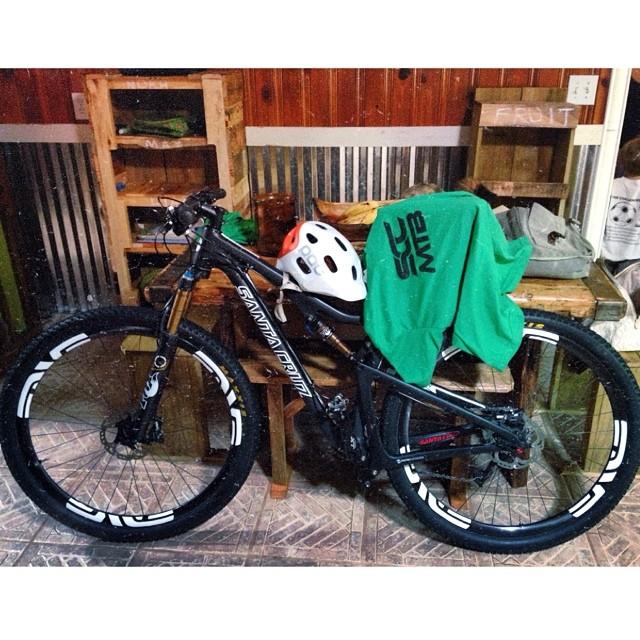 Santa Cruz Tallboy Carbon: A mountain bike resting on a tiled floor next to a wooden table with a green sweatshirt and a white helmet on its handlebars. The background features wooden shelves with items and a metallic wall.