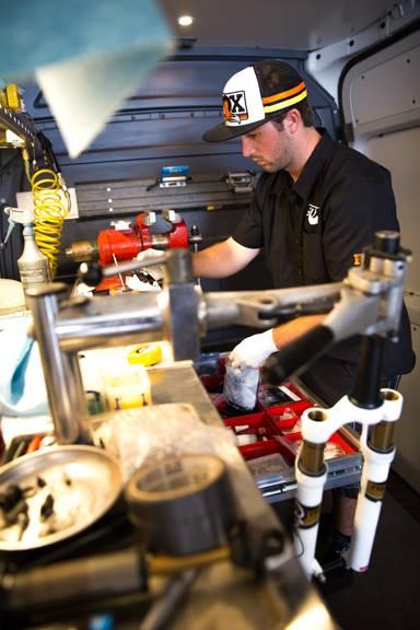A bicycle mechanic works diligently in a mobile workshop, surrounded by various bike tools and equipment. He is focused on inspecting or repairing a bike component, with organized tool trays and a cluttered workspace visible in the backdrop. Keystone Resort Bike Park mountain bike trail.