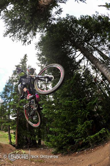 A mountain biker airborne above a dirt trail, surrounded by tall trees and greenery. The bike is lifted in a mid-jump position, showcasing a dynamic riding posture with a focus on the bike's large wheels and the rider's gear. Keystone Resort Bike Park mountain bike trail.
