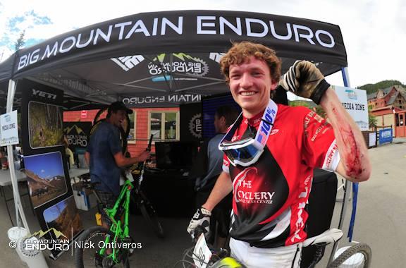 A smiling mountain biker in a red jersey stands in front of a promotional tent for Big Mountain Enduro. He is giving a thumbs-up with one hand while the other hand has visible scrapes, suggesting he has just participated in a race or event. In the background, other cyclists are preparing their bikes. Keystone Resort Bike Park mountain bike trail.