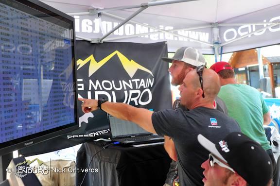 A group of people gathered under a tent at an outdoor event, closely observing a large display screen that shows event-related information. One person, wearing a black shirt and a baseball cap, is pointing at the screen, while others look on with interest. The tent is decorated with the logo of the "Big Mountain Enduro" event. Keystone Resort Bike Park mountain bike trail.