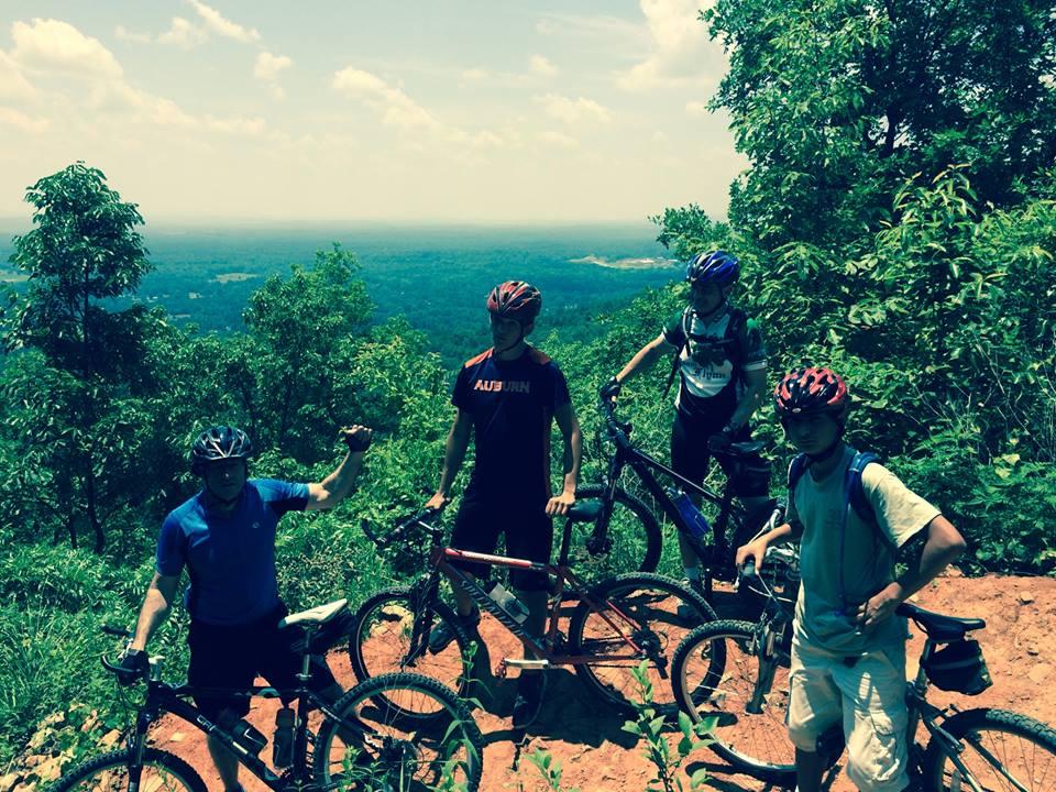 Four mountain bikers standing on a rocky trail overlooking a scenic view of hills and valleys in the background. They are wearing helmets and casual cycling attire, with their bikes beside them. The sky is bright with a few clouds, and lush greenery surrounds the area. Coldwater Mountain mountain bike trail.