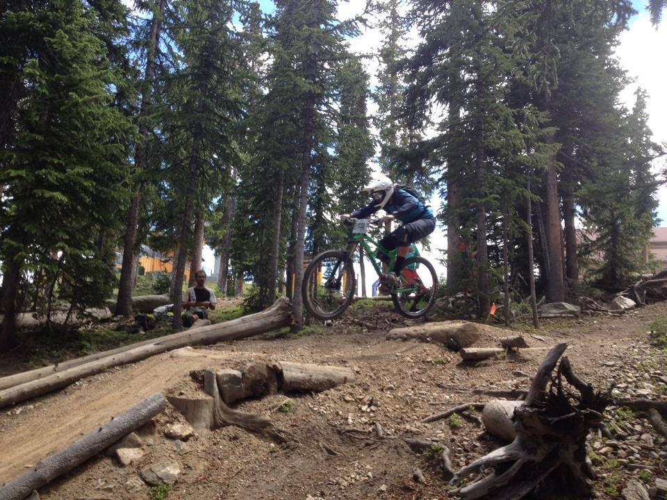 A mountain biker wearing a helmet and protective gear jumps over a log on a forested trail, surrounded by tall trees and rocky terrain. In the background, another person is seated, observing the trail. Bright blue sky is visible through the tree branches. Keystone Resort Bike Park mountain bike trail.