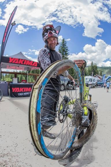 A smiling mountain biker is holding a large bicycle wheel at an outdoor event, with tents and trees visible in the background. The biker wears protective gear and appears cheerful, while the wheel shows signs of use. The sky is partly cloudy, creating a bright and lively atmosphere. Keystone Resort Bike Park mountain bike trail.