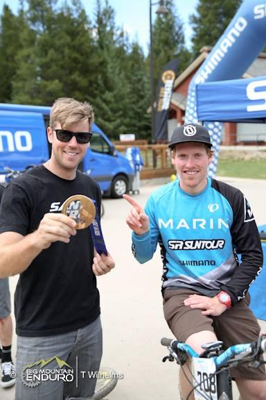 Two men pose for a photo at an outdoor venue, likely after a mountain biking event. One man, wearing a black t-shirt and sunglasses, holds out a medal, while the other, dressed in a blue and black cycling jersey, sits on a mountain bike and smiles. In the background, there are tents and a blue vehicle. The setting appears to be a sunny day in a forested area. Keystone Resort Bike Park mountain bike trail.