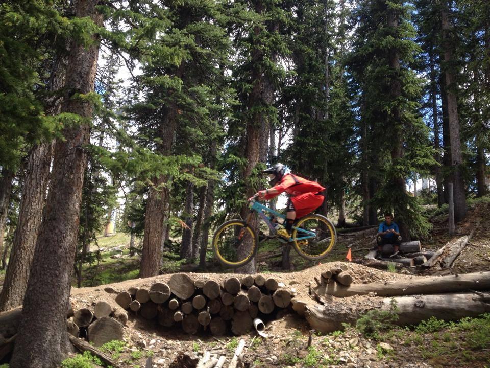 A mountain biker in a red outfit jumps off a dirt ramp in a forested area, surrounded by tall pine trees. Another person sits on a log in the background, watching the action. The scene captures the excitement of outdoor biking in a natural setting. Keystone Resort Bike Park mountain bike trail.