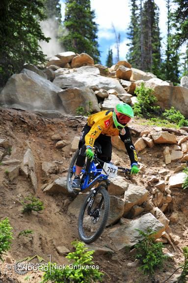A mountain biker in a yellow jersey and green helmet navigates a rocky downhill trail surrounded by trees. Dust kicks up from the bike's tires as the rider descends over large rocks, showcasing an adventurous moment in an enduro mountain biking event. The scene captures the thrill and skill of the sport. Keystone Resort Bike Park mountain bike trail.