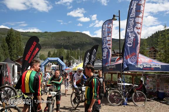 A group of mountain bikers wearing colorful jerseys congregates near vendor tents at an outdoor event. Banners advertising various brands flutter in the breeze, and a colorful inflatable arch marks the entrance to the race area. The background features trees and a clear blue sky, indicating a sunny day in a mountainous setting. Keystone Resort Bike Park mountain bike trail.