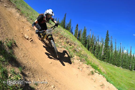A mountain biker navigating a steep, dirt trail on a sunny day, surrounded by lush greenery and pine trees in the background. The rider is wearing safety gear, including a helmet and gloves, and is captured mid-action as they expertly maneuver their bike down the slope. Keystone Resort Bike Park mountain bike trail.