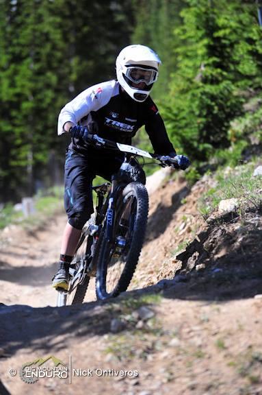 A mountain biker wearing a helmet and protective gear navigates a dirt trail surrounded by trees. The rider leans into the turn, showcasing dynamic movement on a mountain bike in a natural outdoor setting. Keystone Resort Bike Park mountain bike trail.