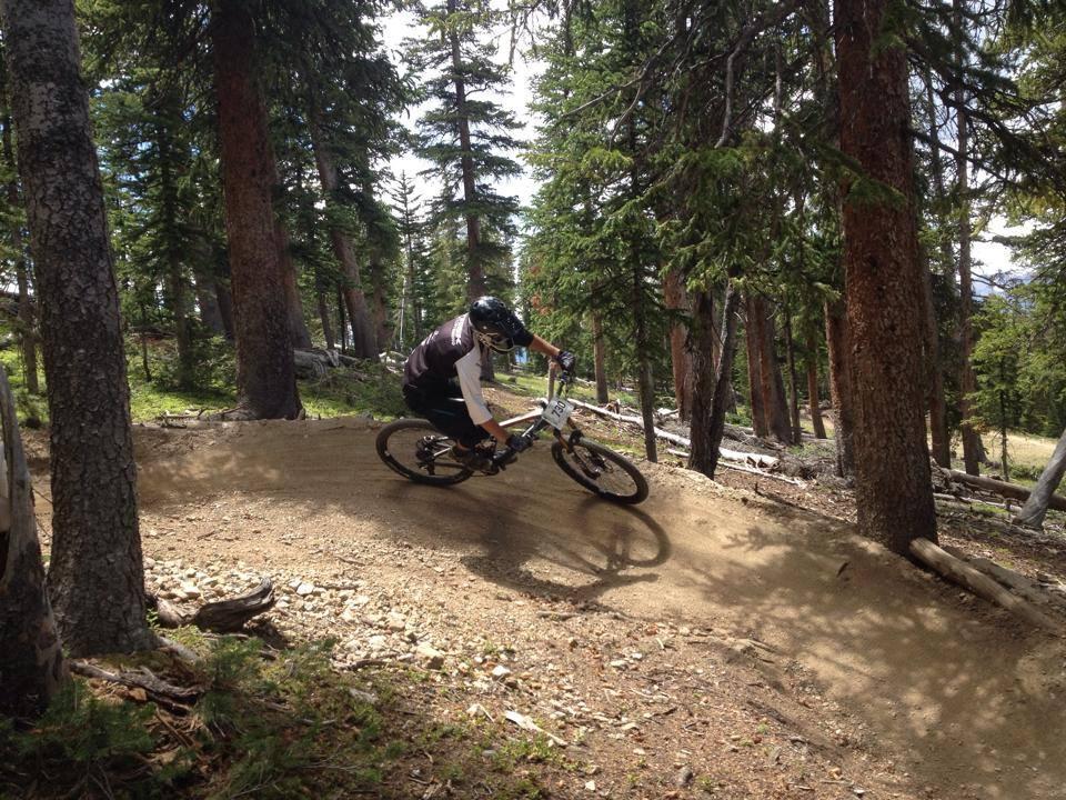 A mountain biker riding on a dirt trail through a forest, leaning into a turn, surrounded by tall trees with green foliage and a partly cloudy sky in the background. Keystone Resort Bike Park mountain bike trail.