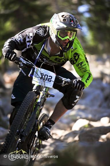A mountain biker in a green and black racing jersey navigates a rocky trail, leaning forward intensely as he rides downhill. He is wearing a helmet with goggles, and his bike features thick tires suited for the rugged terrain. The background shows trees and a blurred landscape indicative of a mountain biking event. The racer wears race number 002. Keystone Resort Bike Park mountain bike trail.