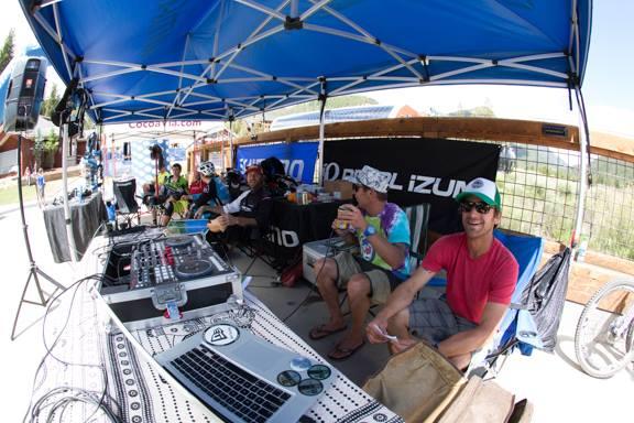 A group of people relaxing under a blue tent at an outdoor event, with a DJ setup visible in the foreground. One person in a colorful shirt and hat is enjoying a drink, while others sit nearby in chairs, engaging in conversation. The backdrop features promotional banners and a sunny outdoor setting. Keystone Resort Bike Park mountain bike trail.