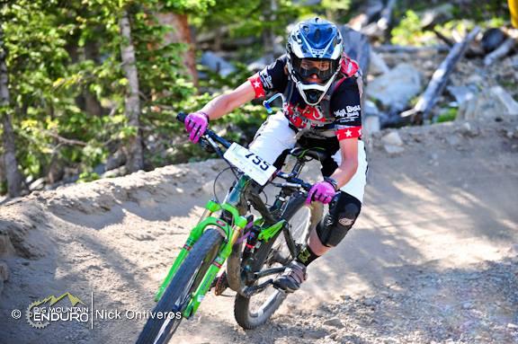 A mountain biker in protective gear races down a dirt trail, leaning into a turn. The cyclist is wearing a helmet with goggles and brightly colored gloves, and their bike is predominantly green. A race number is visible on their jersey. Lush trees and rugged terrain are visible in the background, suggesting a mountain trail setting. Keystone Resort Bike Park mountain bike trail.